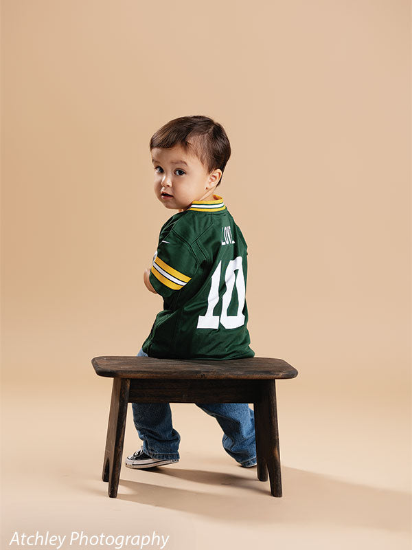 Toddler in a green football jersey with yellow striped sleeves and the number 10 on the back sitting sideways on a rustic wooden bench, looking over his shoulder toward the camera against a warm khaki brown seamless studio backdrop.