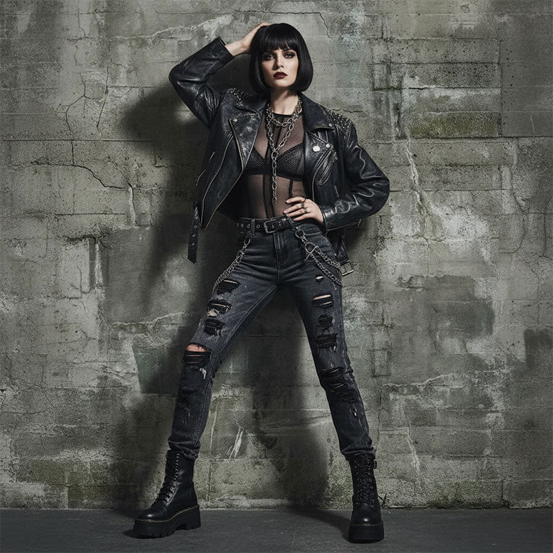 Woman in black leather jacket, mesh top, ripped black jeans, and boots stands with legs apart and one hand on her head in front of a weathered gray concrete block wall backdrop.