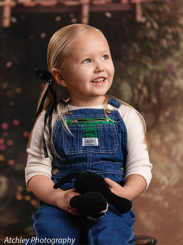 A young girl wearing denim overalls and a white long sleeve shirt sits holding a stuffed animal, smiling gently, with a whimsical forest treehouse garden backdrop filled with greenery and blossoms.
