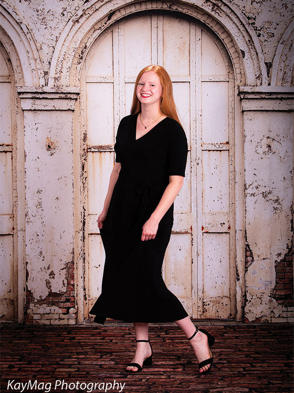 Teen girl in a black dress walking joyfully on an aged brick floordrop in front of a weathered white arched door backdrop, great for elegant and vintage-inspired portraits.