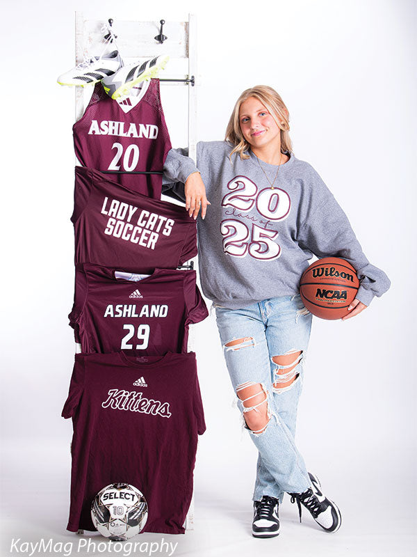High school girl in Class of 2025 sweatshirt posing with sports jerseys, soccer ball, and basketball against a white sports backdrop for senior photos.