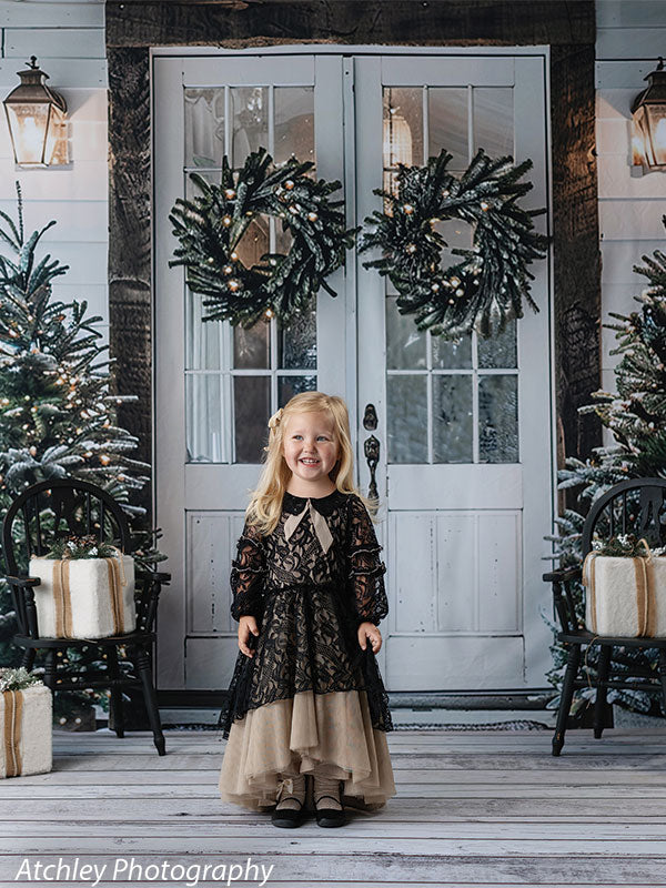 Christmas backdrop with white French doors, holiday wreaths, decorated trees, and presents, shown with a young girl in a black lace dress, ideal for elegant seasonal studio photography.