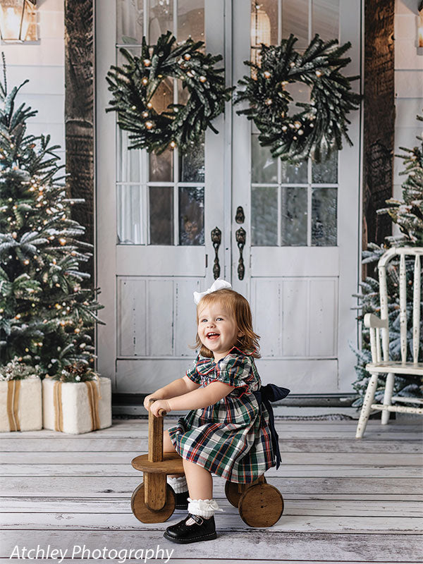 White Christmas doors photography backdrop with evergreen wreaths, snowy trees, and wrapped gifts, featuring a young girl riding a wooden toy, perfect for festive holiday portraits.