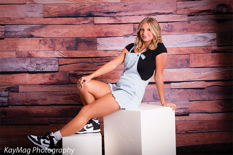 High school senior sitting on a pair of white display cubes in front of a rustic wood backdrop, using cube props for posing in casual lifestyle portraits.