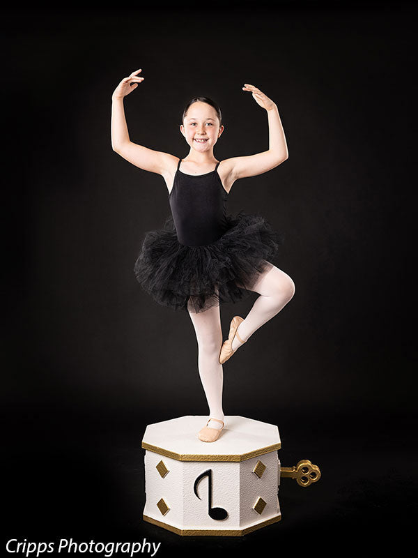 Young ballerina in performance pose balancing on one leg atop a white and gold wind-up music box prop, adding a graceful focal point for ballet-inspired portrait sessions.