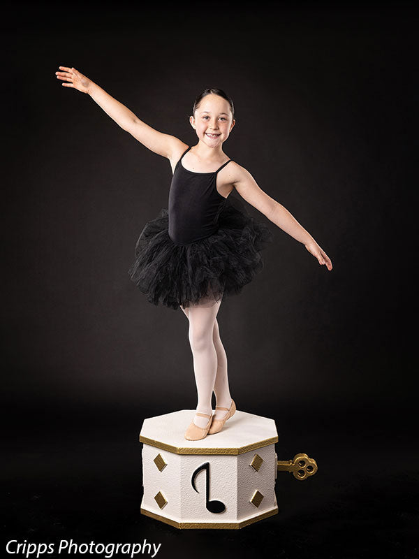 Smiling child ballerina strikes an extended arm pose while standing on a white and gold wind-up music box prop, perfect for dance recital and whimsical themed backdrops.