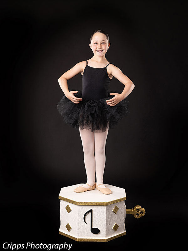 Child ballet dancer in black leotard and tutu poses in first position while standing on a white and gold wind-up music box prop, ideal for dance studio and recital-themed photo setups.