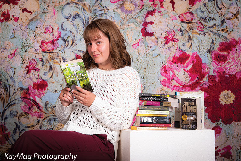 Student seated beside a stack of books on a white cube prop with a vintage floral tapestry backdrop, perfect for literary or themed senior photo sessions.