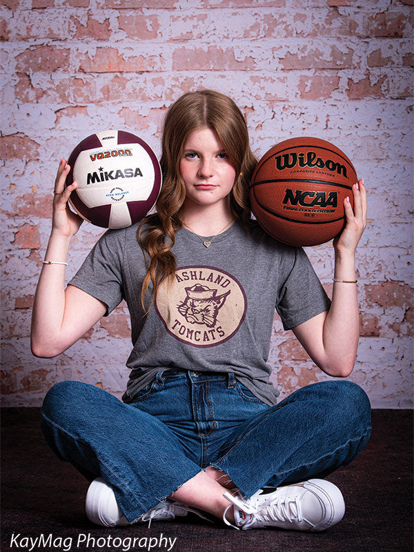 Teen girl seated cross-legged on dark flooring, holding a volleyball and basketball in front of a rustic pink and white brick wall backdrop, great for sporty school-themed photo sessions.