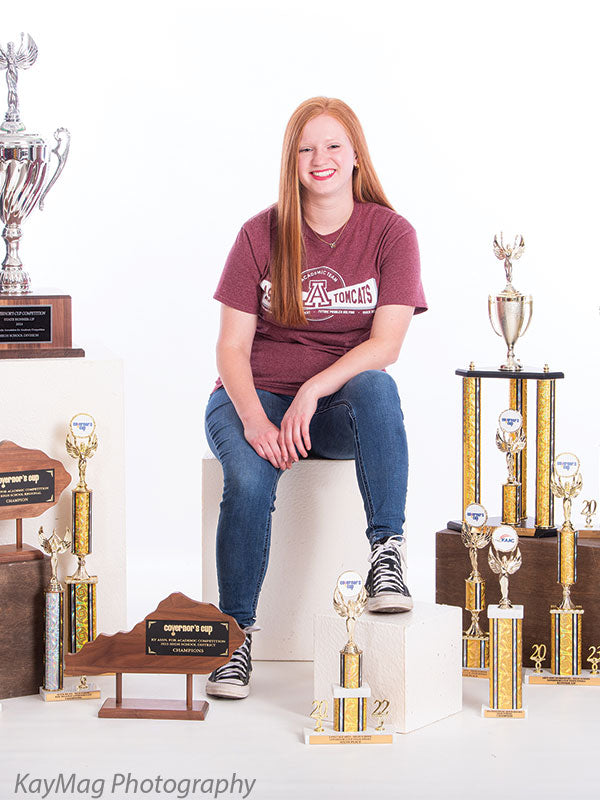 Senior athlete surrounded by sports trophies while sitting on white podium cube props in a clean white studio, perfect for school achievement or championship portraits.
