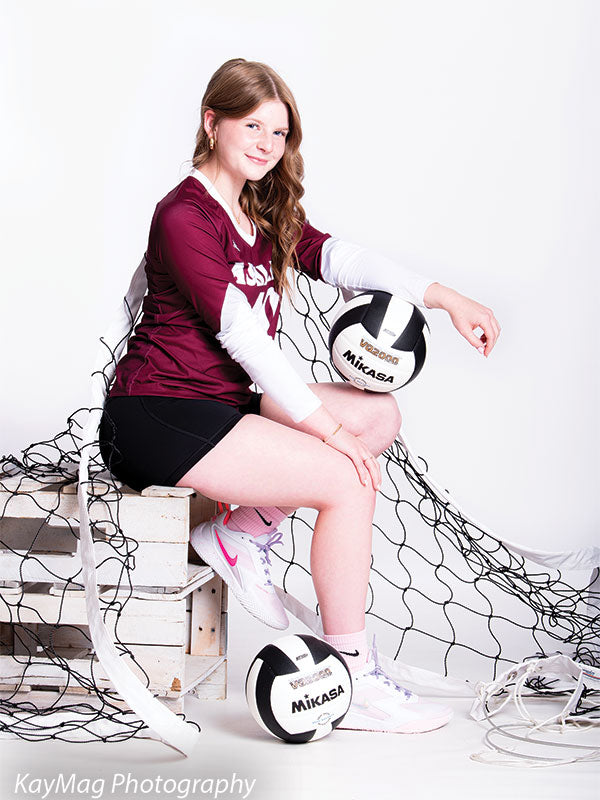 Teen girl in volleyball uniform posing with net and Mikasa volleyballs on a white backdrop, ideal for athlete senior portraits.