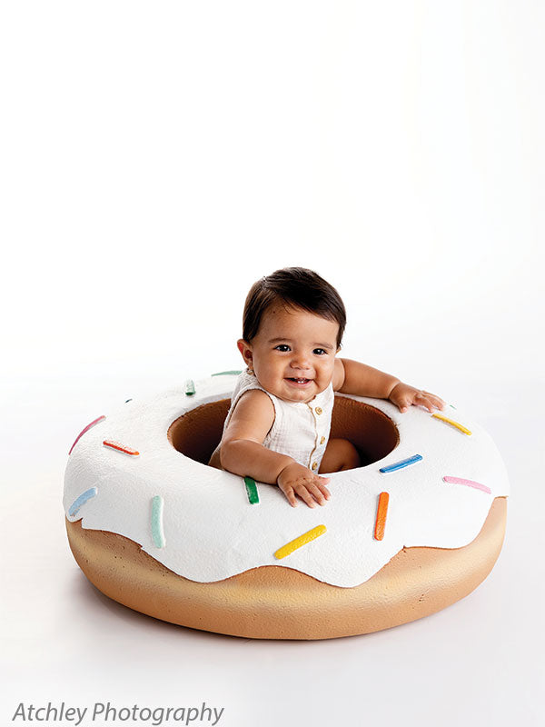 Baby in a cream sleeveless romper sitting inside a large donut prop with white icing and colorful sprinkles, positioned against a white studio background, smiling while leaning forward with hands resting on the donut edge.