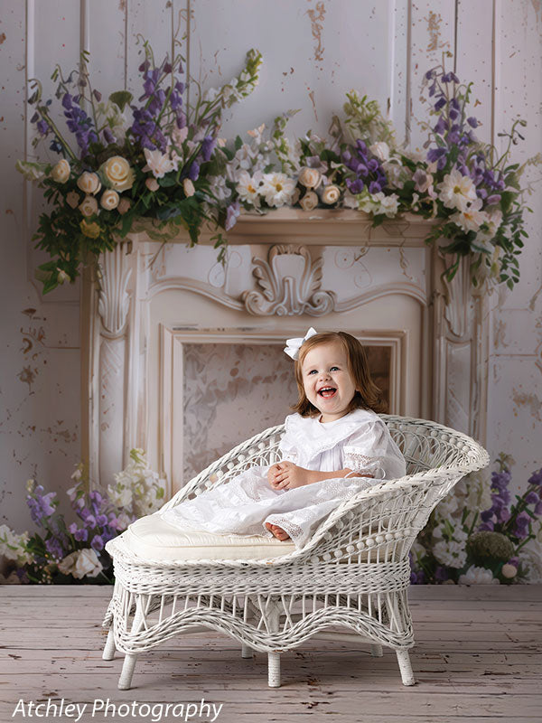 Toddler girl in a white dress with a bow, laughing while sitting on a white wicker chair in front of a vintage floral mantle studio backdrop.
