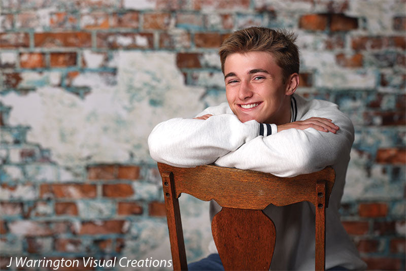 Teen boy smiling over the back of a wooden chair with a whitewashed brick wall photo backdrop, perfect for lifestyle and senior sessions.