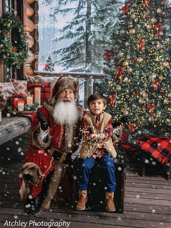 Santa Claus and a young boy seated on a wooden trunk on a log cabin porch as snow falls around them, with a brightly lit Christmas tree decorated in red and gold, wreaths, candles, and snowy pine trees creating a warm winter holiday backdrop.
