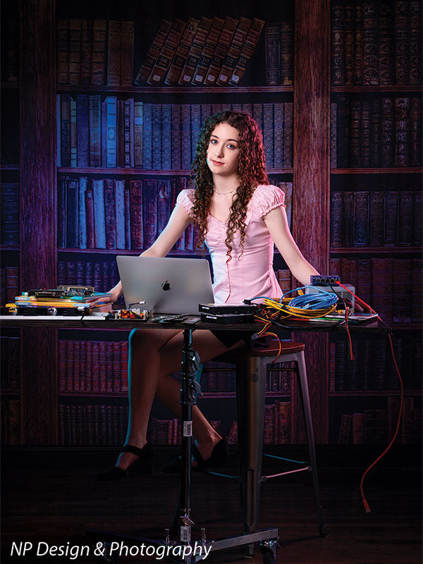 Worn volumes book shelf photography backdrop shown with a senior girl in a pink top seated at a desk with a laptop and colorful cables CP-7317
