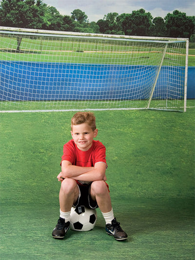 Photography backdrop showing a realistic soccer goal on a wide grassy field with tree-lined scenery.