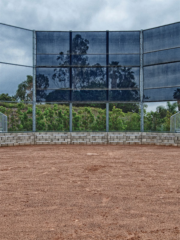 Baseball or Softball Field Photo Backdrop - Softball field with reddish-brown infield, green grass, blue backstop netting, and lush greenery under an overcast sky, perfect for sports-themed photography.