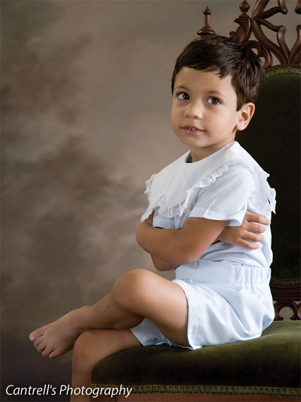 Young boy in white sits on a vintage chair with a painted look nature-inspired backdrop of brown tree silhouettes—ideal for classic boys’ portraits and traditional children’s sessions.