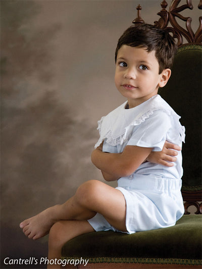 Young boy in white sits on a vintage chair with a painted look nature-inspired backdrop of brown tree silhouettes—ideal for classic boys’ portraits and traditional children’s sessions.