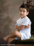 Young boy in white sits on a vintage chair with a painted look nature-inspired backdrop of brown tree silhouettes—ideal for classic boys’ portraits and traditional children’s sessions.