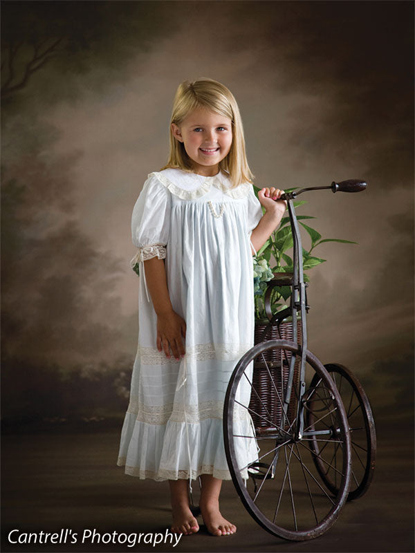 Little girl poses barefoot beside a vintage tricycle in front of a soft-toned floral garden backdrop with painterly trees, perfect for timeless children's portraits and heirloom-style photography.