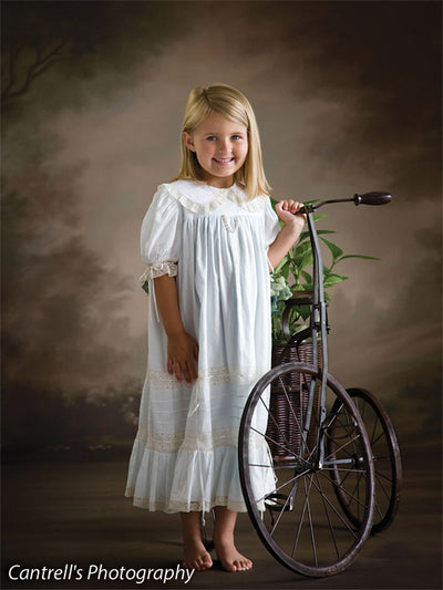 Little girl poses barefoot beside a vintage tricycle in front of a soft-toned floral garden backdrop with painterly trees, perfect for timeless children's portraits and heirloom-style photography.