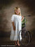 Little girl poses barefoot beside a vintage tricycle in front of a soft-toned floral garden backdrop with painterly trees, perfect for timeless children's portraits and heirloom-style photography.