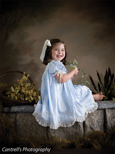 Girl in heirloom blue dress sits on stone ledge in front of a hand-painted floral garden backdrop—ideal for storytelling-style children’s photography and family scenes.