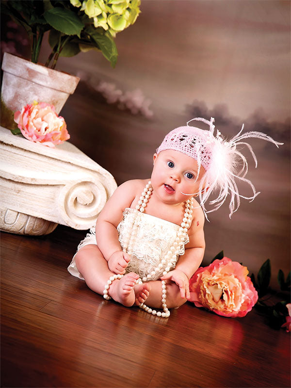 Newborn girl in pearls and feathered headband sits on hardwood floor with vintage column and floral props against a pink-brown painted backdrop—perfect for newborn and baby photography.