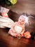 Newborn girl in pearls and feathered headband sits on hardwood floor with vintage column and floral props against a pink-brown painted backdrop—perfect for newborn and baby photography.
