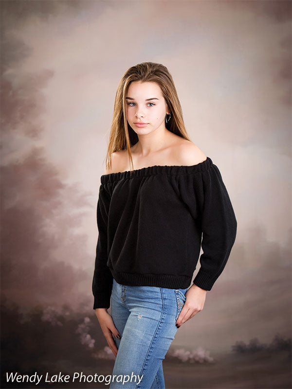 Teen girl poses in front of soft pink and taupe clouded sky backdrop with subtle florals, ideal for senior portraits and styled studio photography.