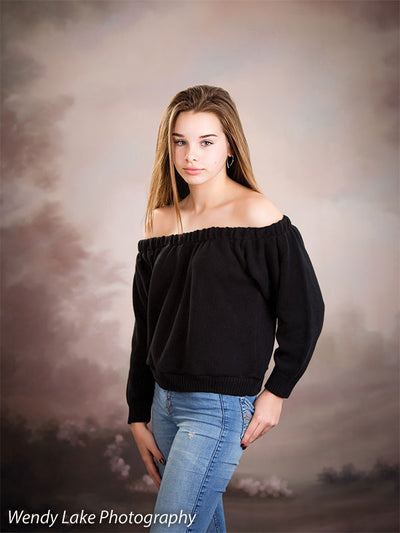 Teen girl poses in front of soft pink and taupe clouded sky backdrop with subtle florals, ideal for senior portraits and styled studio photography.