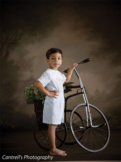 Boy poses with tricycle in front of a romantic garden backdrop painted in warm tones—suited for family, school, or vintage children's portraiture.