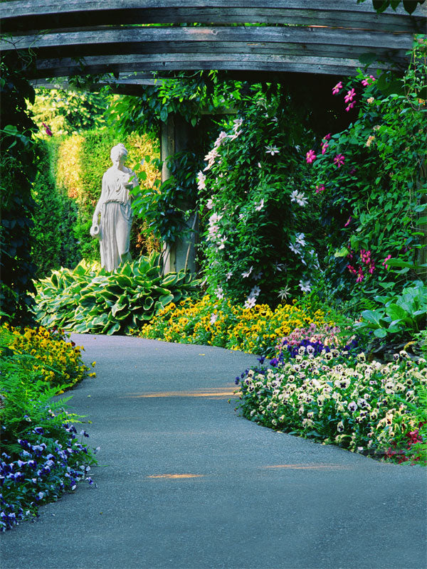 A romantic photography backdrop featuring a winding stone path through vibrant flower beds, lush greenery, and a classical statue beneath a wooden arbor.