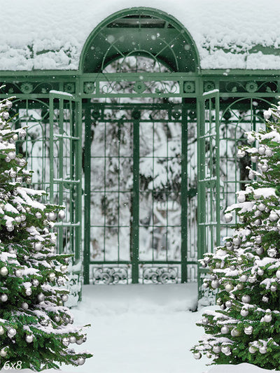 Enchanted Winter Garden Photography Backdrop - Photography backdrop featuring a serene winter garden with a green wrought-iron gate, two snow-covered Christmas trees, and fresh snow.