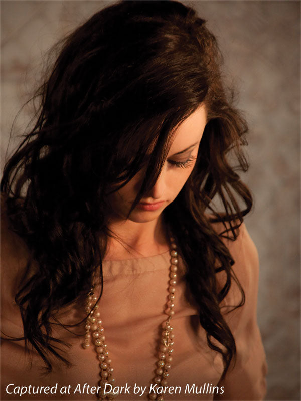 Female model with dark hair poses under warm lighting in front of a soft beige European tile backdrop—perfect for senior portraits and stylized studio photography.