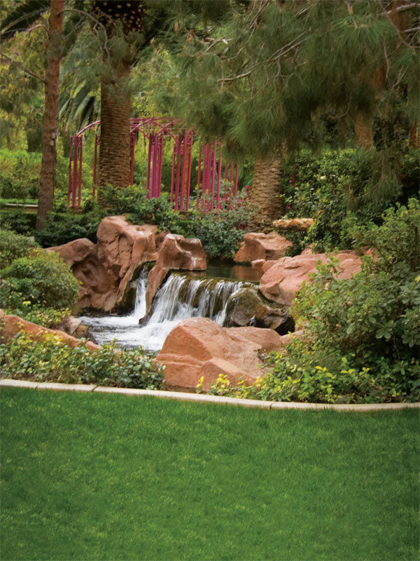Small waterfall flowing over warm red rocks, surrounded by lush greenery and garden elements.