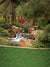 Small waterfall flowing over warm red rocks, surrounded by lush greenery and garden elements.