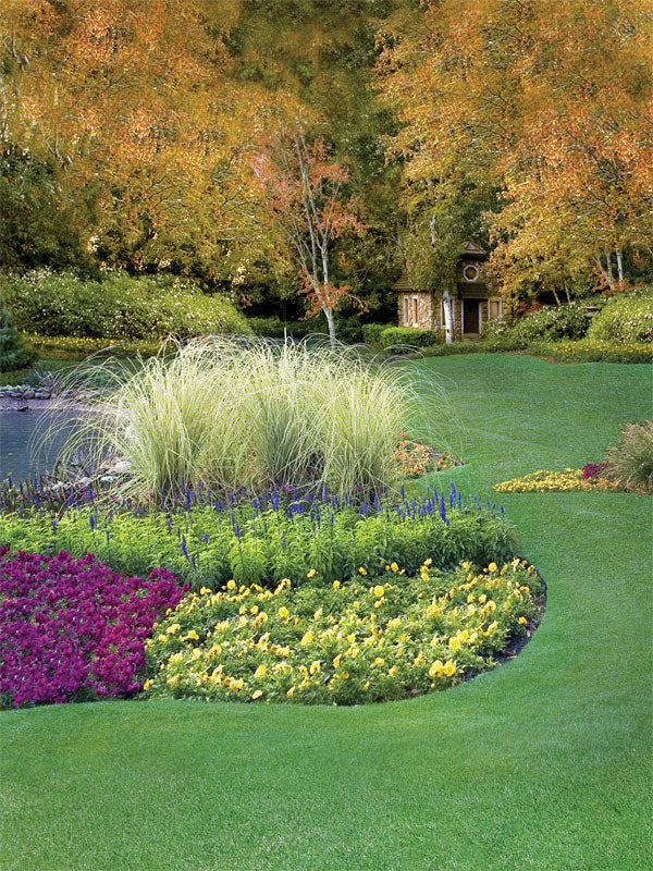 A colorful fall photography backdrop featuring manicured flower beds, ornamental grasses, a pond edge, and a hidden garden cottage under autumn trees.