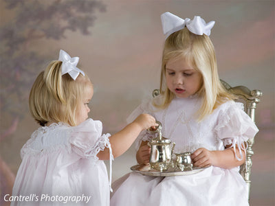 Two girls dressed in white lace gowns host a tea party against a dreamy, floral backdrop—suitable for vintage-style children’s portraits.