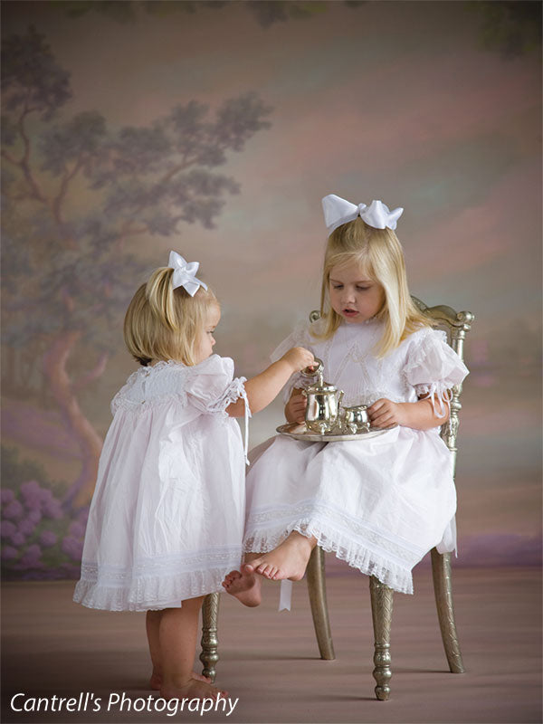 Close-up of girls in heirloom dresses playing tea party with a soft, pastel floral scene—perfect for elegant and detailed children’s photography.