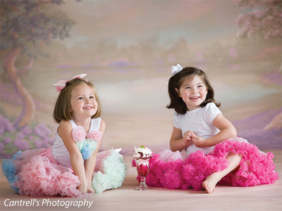 Two young girls in pastel tutus sit cheerfully in front of a soft pastel garden backdrop—perfect for whimsical children’s portraits and springtime photography.
