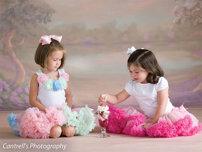 Two young girls dressed in pink and white ruffled skirts sit on a floor in front of a pastel Sheffield garden backdrop, ideal for spring-themed portrait and dance photography.