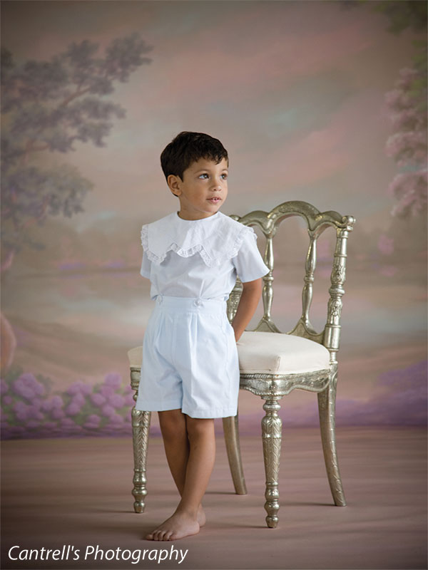 Young boy in heirloom outfit standing with ornate chair in front of pastel floral backdrop—perfect for vintage portraiture and family photography.
