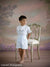 Young boy in heirloom outfit standing with ornate chair in front of pastel floral backdrop—perfect for vintage portraiture and family photography.