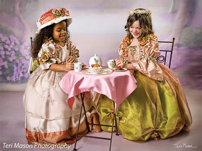 Two girls in elegant Victorian-style gowns enjoy a tea party in front of a delicate floral Sheffield backdrop, great for themed portrait photography and children’s fantasy shoots.