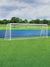 A vibrant green outdoor soccer field backdrop with a regulation goal and overcast sky.
