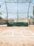 Softball or Baseball Home Plate Backdrop - Home base area of a baseball or softball field with infield dirt, green backstop padding, chain-link fencing, and clear blue sky.