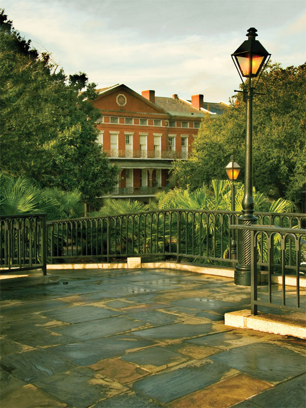 A photography backdrop featuring a historic brick building, lush foliage, and gas lamps in a Southern courtyard setting.